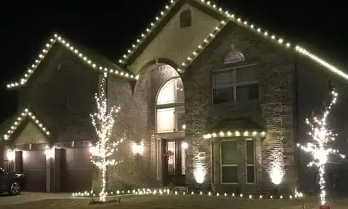 Brick home with warm white roofline lights and lit trees in the front yard